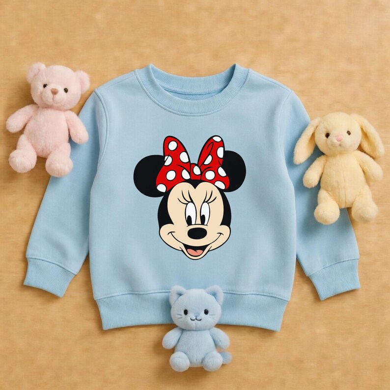 Może przedstawiać: Jasnoniebieska bluza z nadrukiem Myszki Minnie, z czerwoną kokardą w białe grochy. Na zdjęciu znajduje się r&oacute;wnież r&oacute;żowy miś, ż&oacute;łty kr&oacute;lik i niebieska pluszowa zabawka w kształcie kota.