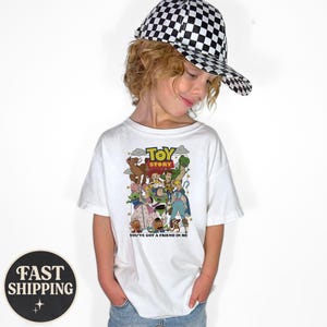 Puede incluir: Camiseta blanca con un gr&aacute;fico colorido de los personajes de Toy Story, el texto "Toy Story" y "You've Got a Friend in Me." La persona lleva una gorra de b&eacute;isbol a cuadros blancos y negros.