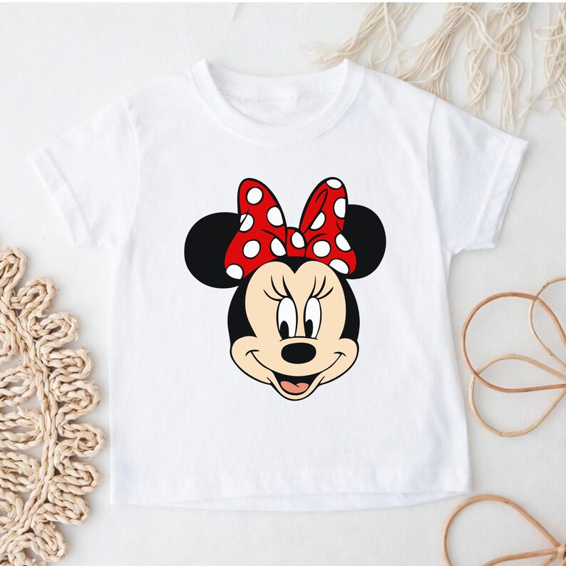 Może przedstawiać: Biała koszulka z motywem Myszki Minnie. Grafika przedstawia twarz Myszki Minnie z czerwoną kokardą w białe grochy. Koszulka ma okrągły dekolt i kr&oacute;tkie rękawy.