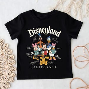 May include: Black t-shirt featuring the word "Disneyland" in a white script font, along with cartoon characters like Mickey Mouse, Minnie Mouse, and Donald Duck. The shirt also includes the text "California" and "EST. 1955".