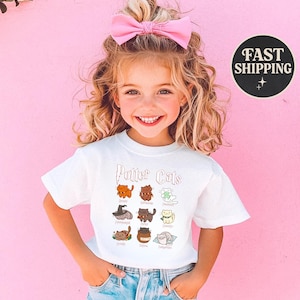 May include: White t-shirt with the text "Potter Cats" and cartoon cat illustrations. The shirt features various cat designs, each with a unique style and color, on a pink background. The child is wearing a pink bow.