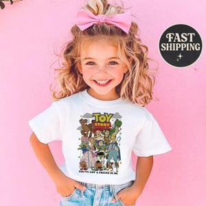 Puede incluir: Una camiseta blanca con un gr&aacute;fico colorido de personajes de la pel&iacute;cula animada "Toy Story". La camiseta incluye el texto "TOY STORY" y "YOU'VE GOT A FRIEND IN ME". La imagen muestra a una ni&ntilde;a peque&ntilde;a usando la camiseta.