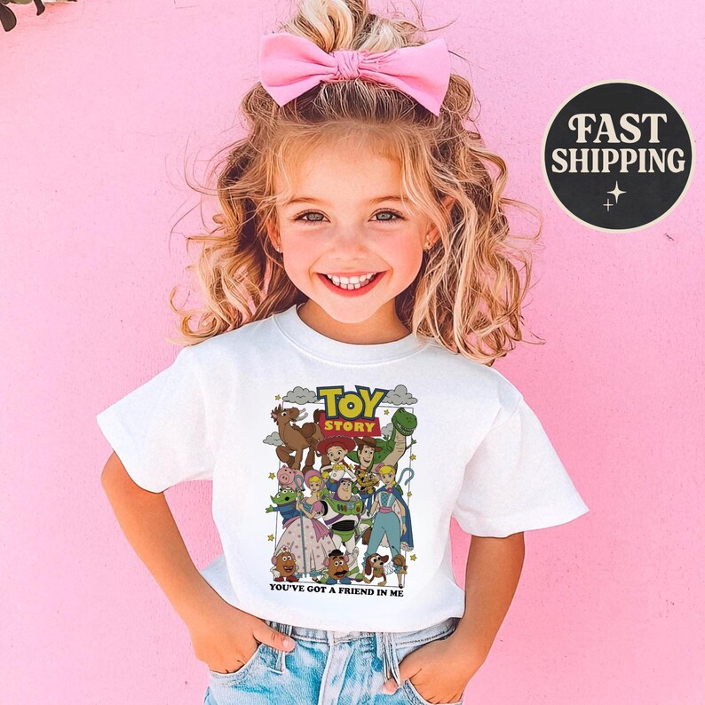 Puede incluir: Una camiseta blanca con un gr&aacute;fico colorido de personajes de la pel&iacute;cula animada "Toy Story". La camiseta incluye el texto "TOY STORY" y "YOU'VE GOT A FRIEND IN ME". La imagen muestra a una ni&ntilde;a peque&ntilde;a usando la camiseta.