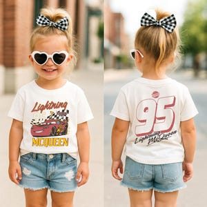 May include: A child wearing a white t-shirt with a Lightning McQueen graphic and the number 95. The shirt is paired with denim shorts and a black and white checkered bow. The child is also wearing heart-shaped sunglasses.
