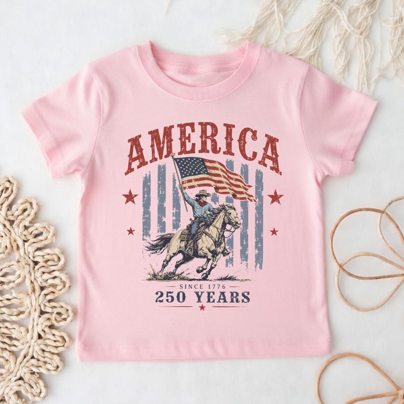 May include: A light pink short-sleeve t-shirt featuring a graphic of a cowboy riding a horse while holding an American flag. The text reads AMERICA at the top and SINCE 1776 250 YEARS at the bottom, surrounded by red stars and a faded flag design.