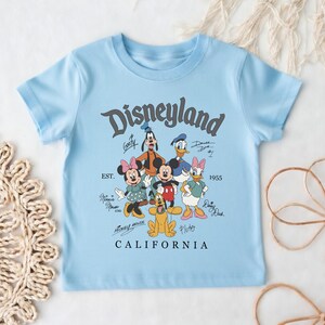 May include: Light blue t-shirt featuring the word "Disneyland" in a vintage font, along with cartoon characters like Mickey Mouse, Minnie Mouse, and Donald Duck. The shirt also includes the text "EST. 1955" and "CALIFORNIA".