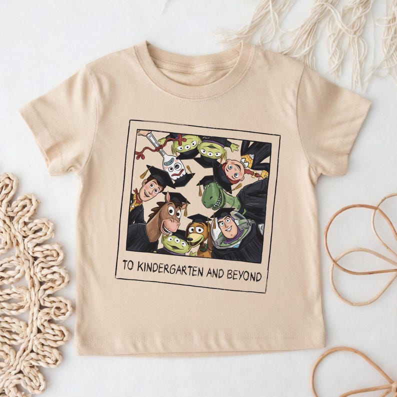 May include: A beige short-sleeve t-shirt featuring a graphic print of Toy Story characters wearing graduation caps. The design is framed like a polaroid photo with the text TO KINDERGARTEN AND BEYOND printed in black at the bottom of the frame.