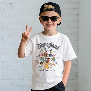 May include: A white t-shirt featuring a Disneyland graphic with Mickey Mouse, Minnie Mouse, Donald Duck, Daisy Duck, Goofy, and Pluto. The shirt has the text "Disneyland California" and "EST. 1955". The person is wearing a black cap and sunglasses.