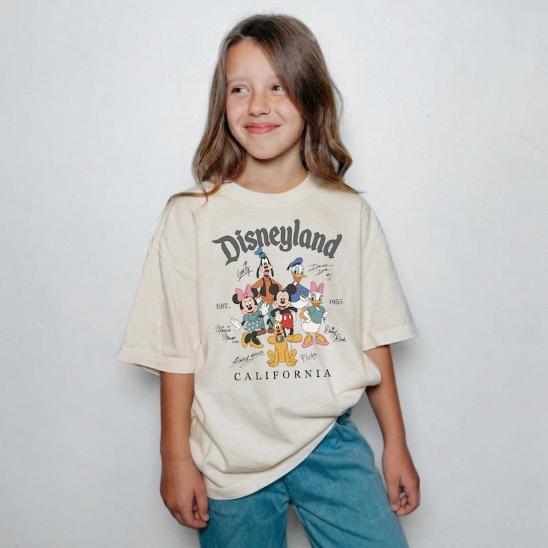 May include: Cream-colored Disneyland t-shirt featuring a graphic of Mickey Mouse, Minnie Mouse, Donald Duck, Daisy Duck, Goofy, and Pluto. The shirt has the text "Disneyland California" in a vintage font.
