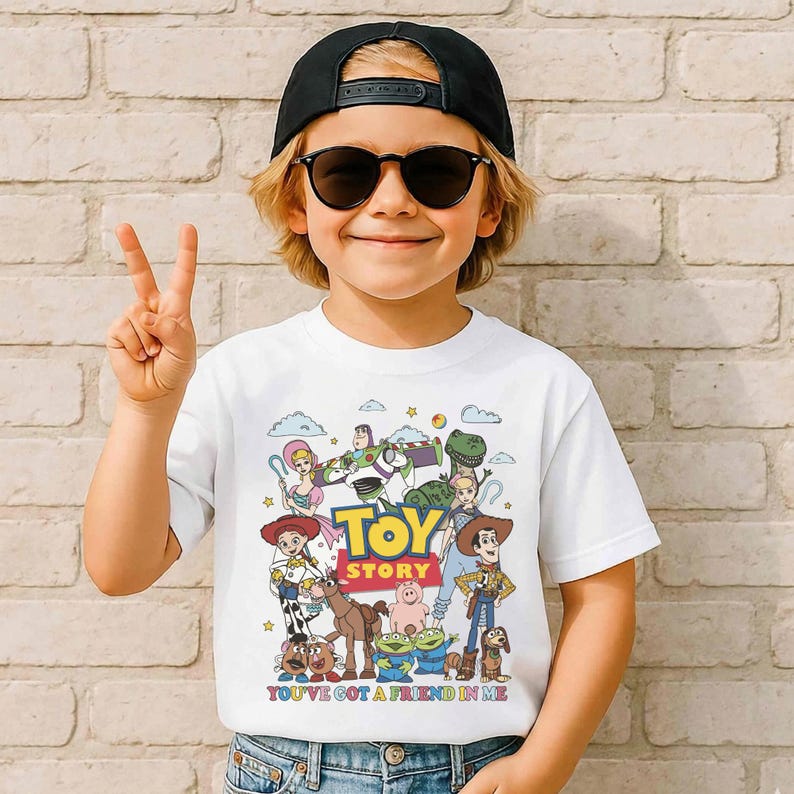 Op de afbeelding: Wit T-shirt met een kleurrijke afbeelding van personages uit de animatiefilm "Toy Story". Het ontwerp bevat de tekst "Toy Story" en "You've Got a Friend in Me". Het shirt wordt gedragen door een persoon die een zwarte pet en zonnebril draagt.