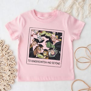 May include: A light pink short-sleeved t-shirt featuring a square graphic of Toy Story characters wearing graduation caps. The print includes the text TO KINDERGARTEN AND BEYOND below the characters, which include Woody, Buzz Lightyear, and Forky.