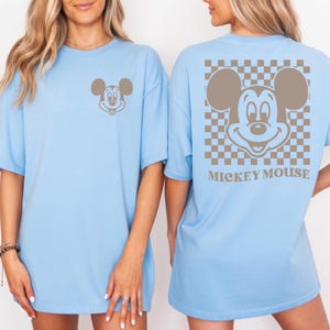 May include: Light blue t-shirt featuring a Mickey Mouse graphic. The front of the shirt has a small Mickey Mouse design, while the back showcases a larger Mickey Mouse design within a brown and white checkered pattern. The words "MICKEY MOUSE" are printed below the design.