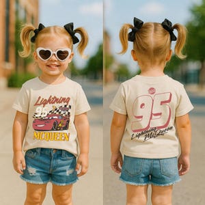 May include: A child wearing a tan t-shirt featuring Lightning McQueen from the Cars movie. The front of the shirt displays the character and text, while the back shows a large "95" and "Lightning McQueen". The child is also wearing denim shorts.