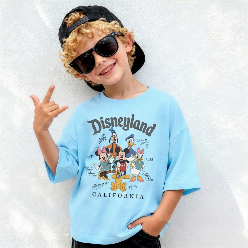 May include: A light blue t-shirt with the word "Disneyland" and cartoon characters. The shirt has short sleeves and the text "CALIFORNIA" at the bottom. The shirt is worn by a person wearing sunglasses and a black cap.
