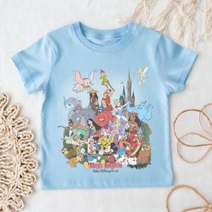 May include: Light blue t-shirt featuring a colorful graphic of various Disney characters, including Dumbo, Peter Pan, and Mickey Mouse. The text "Magic Kingdom, Walt Disney World" is printed at the bottom of the graphic. The shirt is made of a soft material.