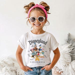 May include: A white t-shirt with a Disneyland graphic featuring Mickey Mouse, Minnie Mouse, Donald Duck, Goofy, and Daisy Duck. The shirt has rolled-up sleeves and the text "Disneyland California 1955". The child is wearing sunglasses and a pink headband.
