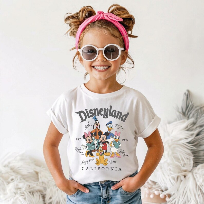 May include: A white t-shirt with a Disneyland graphic featuring Mickey Mouse, Minnie Mouse, Donald Duck, Goofy, and Daisy Duck. The shirt has rolled-up sleeves and the text "Disneyland California 1955". The child is wearing sunglasses and a pink headband.