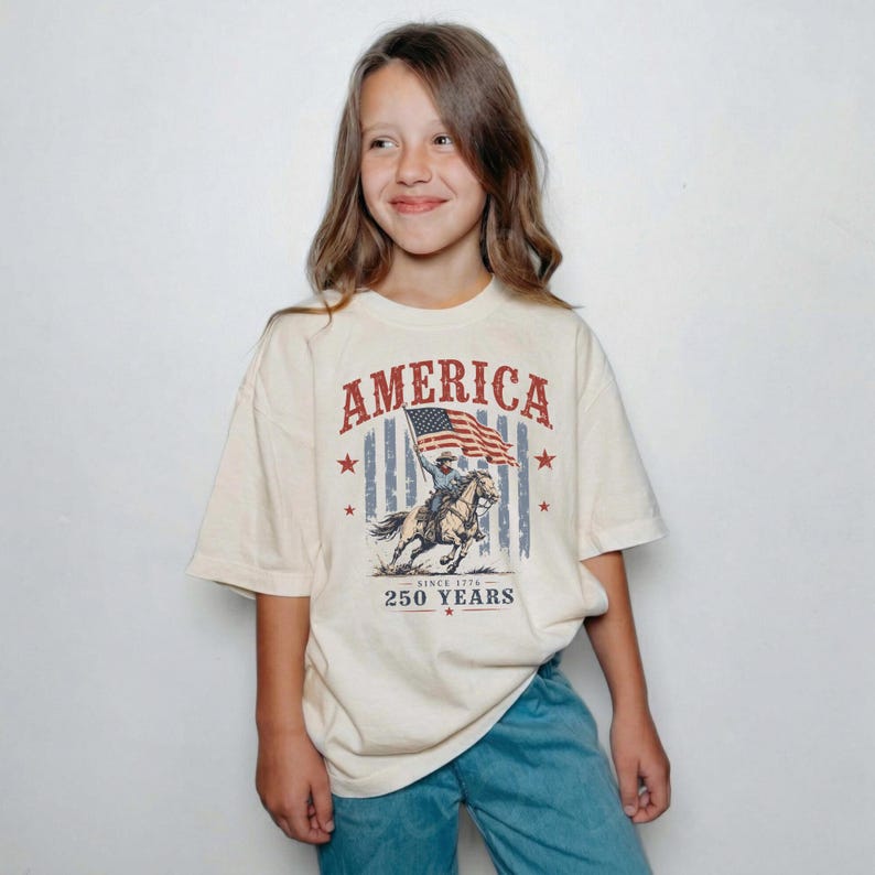 May include: A cream-colored short-sleeve t-shirt featuring a graphic print with the text AMERICA and 250 YEARS. The design includes a cowboy riding a horse while holding an American flag, set against a background of vertical blue stripes and red stars.