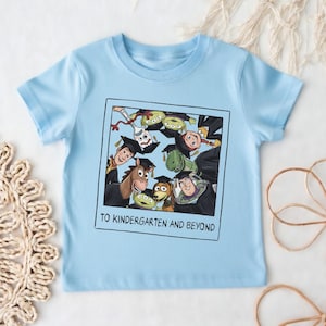 May include: A light blue short-sleeve t-shirt featuring a graphic of Toy Story characters wearing graduation caps inside a square frame. The text below the characters reads TO KINDERGARTEN AND BEYOND in black capital letters.