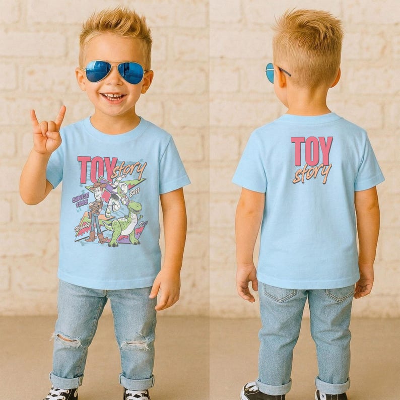 May include: Light blue t-shirt featuring the words "Toy Story" in pink and orange, with a graphic of characters from the movie. The back of the shirt also has the words "Toy Story" in pink and orange. The shirt is worn by a young child.