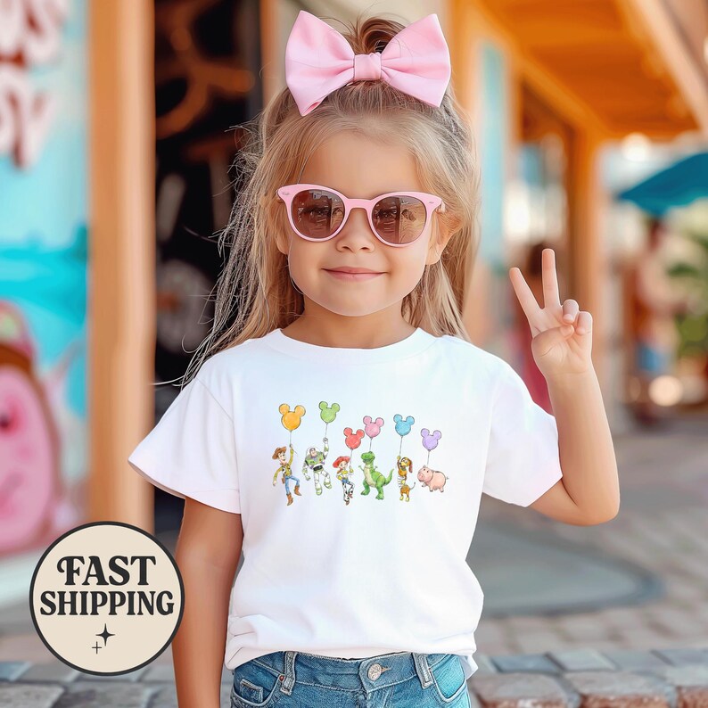 K&ouml;nnte beinhalten: Ein junges M&auml;dchen tr&auml;gt ein wei&szlig;es T-Shirt mit einer bunten Grafik von Zeichentrickfiguren, die ballonf&ouml;rmige Ballons halten. Sie tr&auml;gt eine rosa Sonnenbrille und eine rosa Schleife im Haar. Das Bild enth&auml;lt ein "FAST SHIPPING"-Logo.