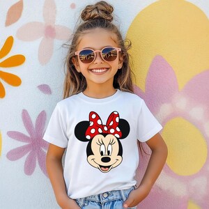 Może przedstawiać: Biała koszulka z grafiką Myszki Minnie. Grafika przedstawia twarz Myszki Minnie, czarne uszy i czerwoną kokardę w białe grochy. Koszulka jest noszona przez osobę noszącą r&oacute;żowe okulary przeciwsłoneczne.