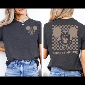 May include: Dark gray t-shirt featuring a tan Mickey Mouse graphic on the front and back. The back design includes a checkered pattern and the words "Mickey Mouse". The shirt is a casual style.