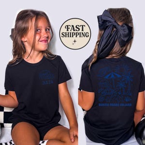 Op de afbeelding: Zwart t-shirt met een blauw golfontwerp en de naam "Julia" op de voorkant. De achterkant van het shirt heeft een blauwe illustratie van een strandsc&egrave;ne met de tekst "South Padre Island".