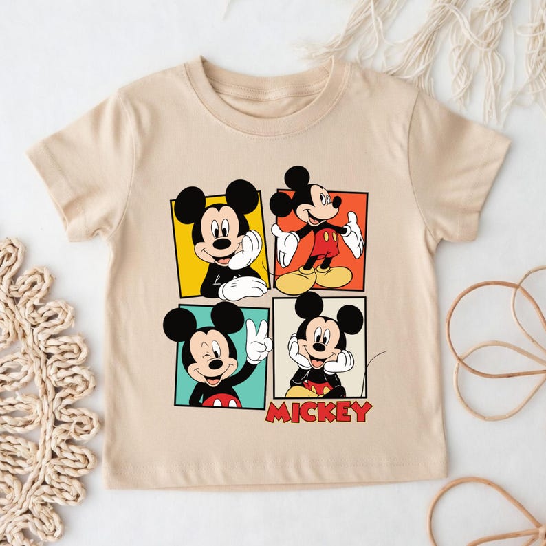May include: A beige short-sleeved t-shirt with a colourful graphic of Mickey Mouse in four different poses. The word "MICKEY" is printed in red below the images. The shirt is made of a soft, comfortable material.