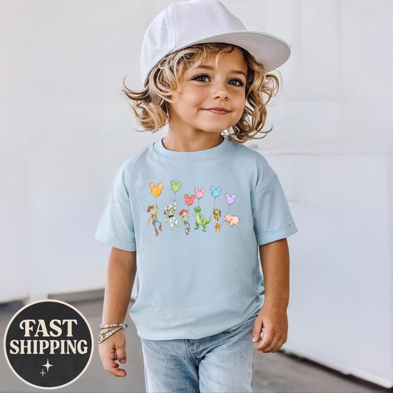 K&ouml;nnte beinhalten: Hellblaues T-Shirt mit einem Cartoon von Toy Story-Charakteren, die Luftballons in Form von Mickey Mouse-K&ouml;pfen halten. Das T-Shirt hat einen Rundhalsausschnitt und kurze &Auml;rmel. Die Worte "FAST SHIPPING" sind auf einem schwarzen Kreis aufgedruckt.