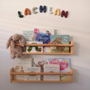 May include: Two light-coloured wooden wall-mounted bookshelves holding various children's books and a plush brown rabbit toy. Above the shelves, the name LACHLAN is spelled out in colourful, rounded, three-dimensional letters on the wall.