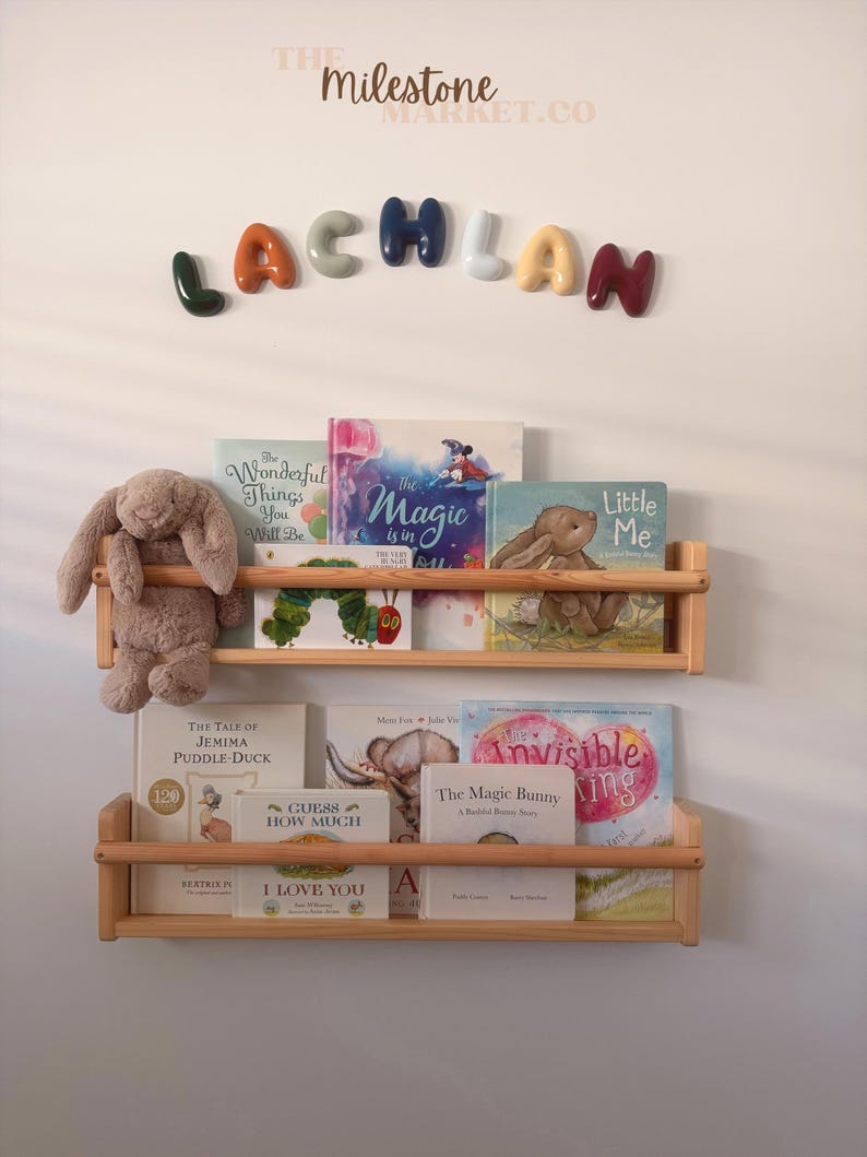 May include: Two light-coloured wooden wall-mounted bookshelves holding various children's books and a plush brown rabbit toy. Above the shelves, the name LACHLAN is spelled out in colourful, rounded, three-dimensional letters on the wall.