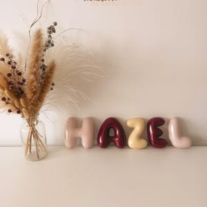 May include: A clear glass vase containing a bouquet of dried pampas grass and small dark berries, positioned next to five individual balloon-style letters spelling the name HAZEL in alternating shades of light pink, dark red, and cream.