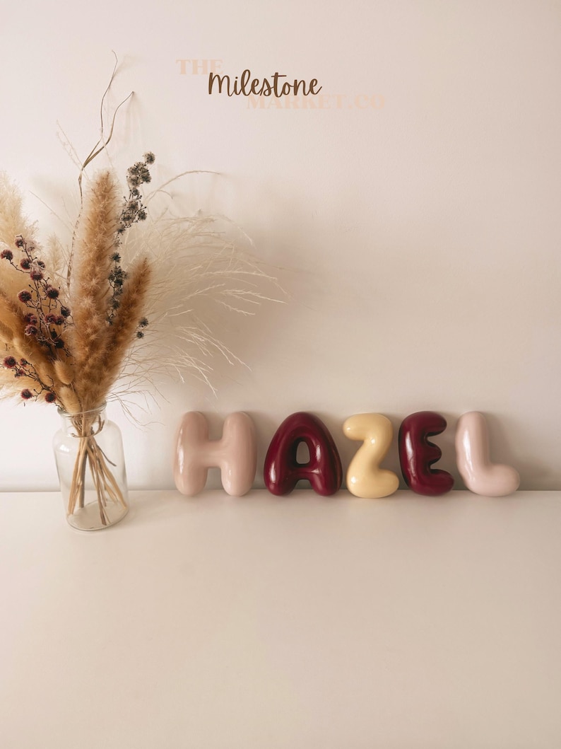 May include: A clear glass vase containing a bouquet of dried pampas grass and small dark berries, positioned next to five individual balloon-style letters spelling the name HAZEL in alternating shades of light pink, dark red, and cream.