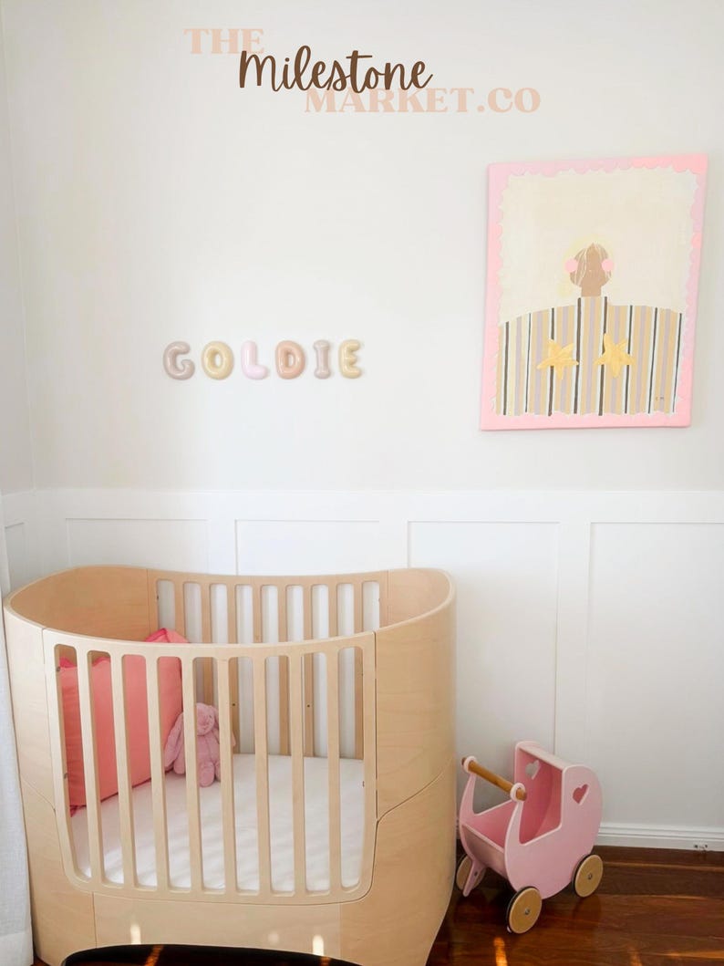 May include: A light wood oval cot with a white mattress, a coral cushion, and a pink plush toy. Beside it sits a small pink wooden toy pram. On the wall, the name GOLDIE is spelled out in letters, and a framed painting shows a figure in a striped shirt.