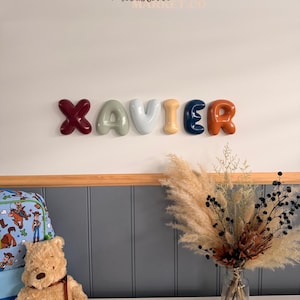 May include: A wall-mounted name sign spelling XAVIER in individual, glossy, balloon-style letters. The letters are coloured dark red, sage green, light grey, mustard yellow, navy blue, and burnt orange. The text THE MILESTONE MARKET.CO is visible.