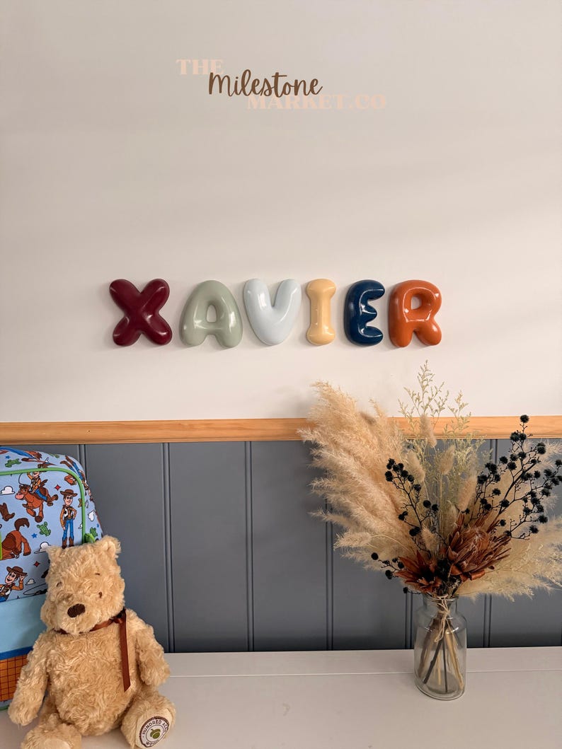 May include: A wall-mounted name sign spelling XAVIER in individual, glossy, balloon-style letters. The letters are coloured dark red, sage green, light grey, mustard yellow, navy blue, and burnt orange. The text THE MILESTONE MARKET.CO is visible.