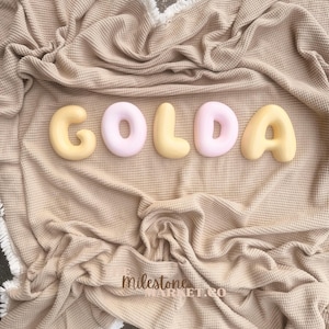 May include: A beige waffle-textured blanket with white fringe edges features five rounded, three-dimensional letters spelling GOLDA in alternating pale yellow and soft pink colours. The text Milestone MARKET.CO is visible in the lower centre.