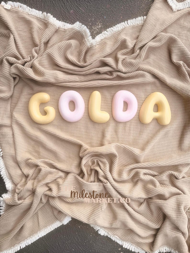 May include: A beige waffle-textured blanket with white fringe edges features five rounded, three-dimensional letters spelling GOLDA in alternating pale yellow and soft pink colours. The text Milestone MARKET.CO is visible in the lower centre.