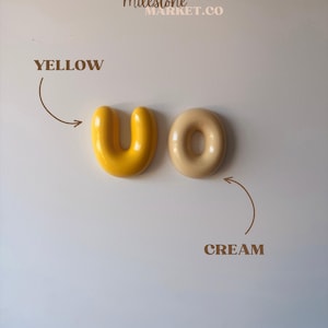 May include: Two smooth, rounded ceramic objects, one U-shaped and bright yellow, the other O-shaped and cream-coloured. Text reads THE MILESTONE MARKET.CO, YELLOW, and CREAM with arrows pointing to each object, indicating their respective colour names.