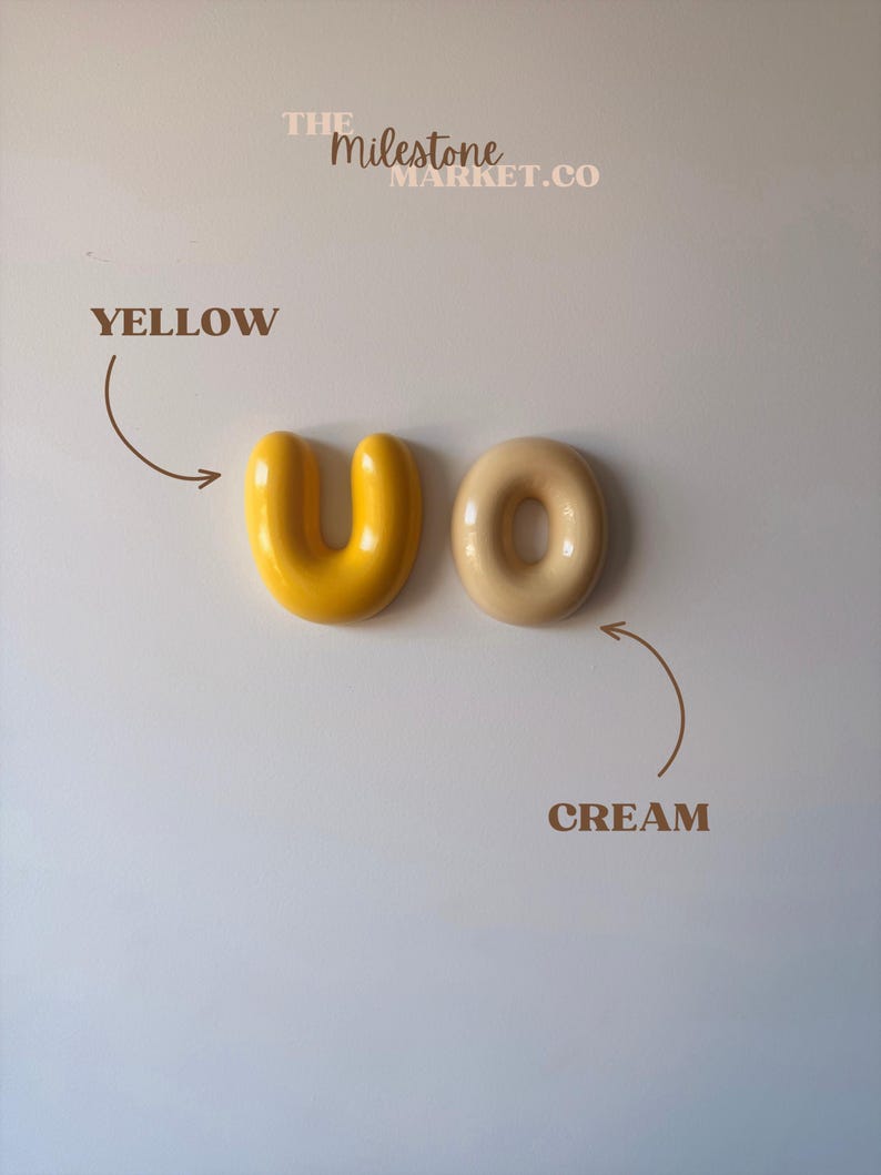 May include: Two smooth, rounded ceramic objects, one U-shaped and bright yellow, the other O-shaped and cream-coloured. Text reads THE MILESTONE MARKET.CO, YELLOW, and CREAM with arrows pointing to each object, indicating their respective colour names.