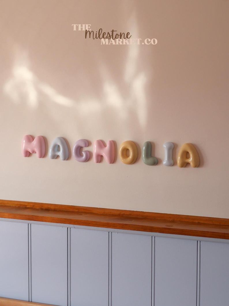 May include: A set of individual, rounded, three-dimensional letters spelling the word MAGNOLIA in various pastel colours including pink, blue, purple, yellow, and green. The text THE MILESTONE MARKET.CO is printed in white above the letters.