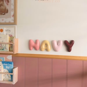 May include: Four individual, rounded, balloon-style wall letters spelling the word NAVY. The letters are coloured in shades of coral, yellow, light pink, and deep maroon, mounted horizontally on a white wall above a pink panelled wainscoting.