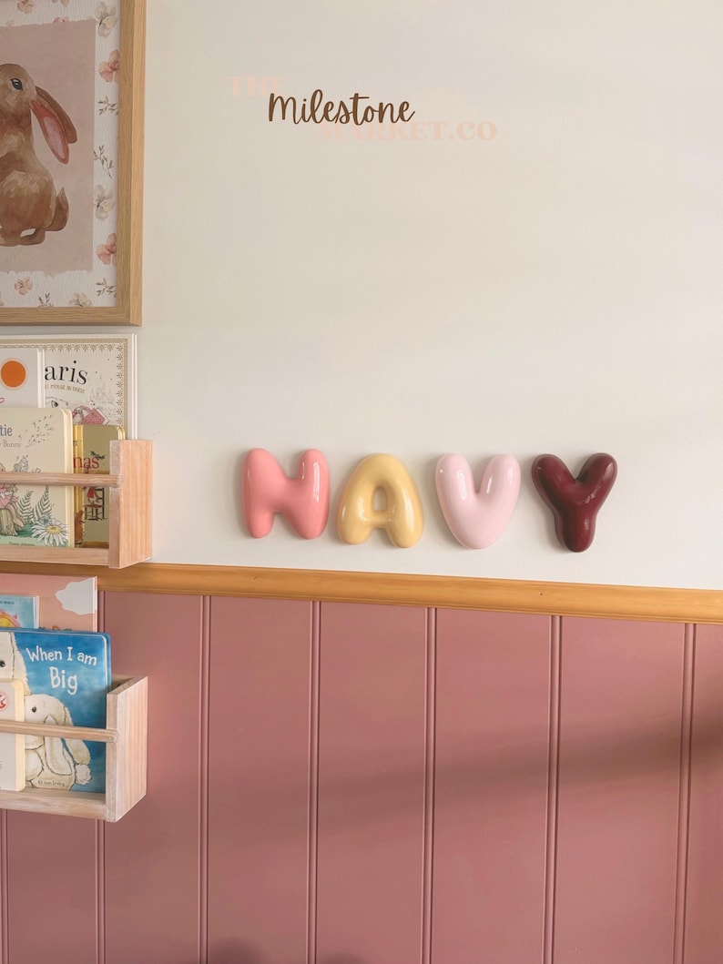 May include: Four individual, rounded, balloon-style wall letters spelling the word NAVY. The letters are coloured in shades of coral, yellow, light pink, and deep maroon, mounted horizontally on a white wall above a pink panelled wainscoting.