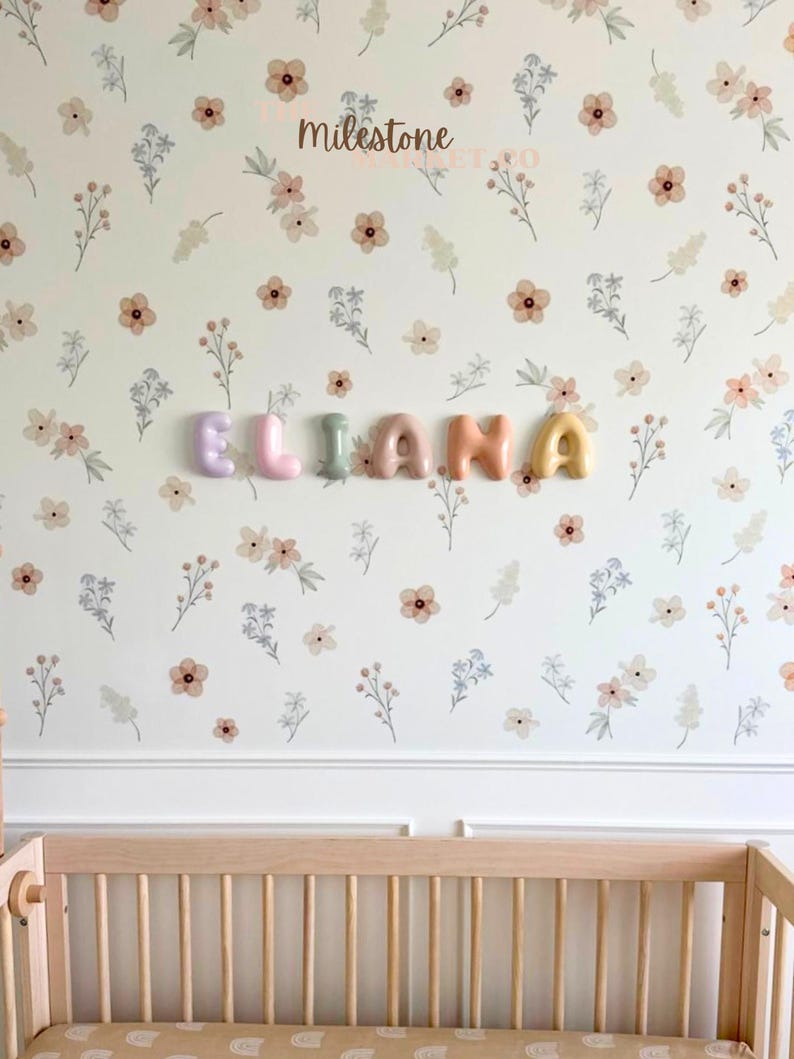 May include: A wall display featuring the name ELIANA spelled out in individual, rounded, pastel-coloured 3D letters. The letters are mounted on a white wall covered in a repeating pattern of small, delicate peach and blue wildflowers.