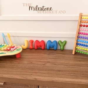 May include: A set of colourful, rounded, three-dimensional letters spelling the name JIMMY, positioned between a wooden xylophone with two mallets and a vertical abacus with rows of multi-coloured beads. The text THE MILESTONE MARKET.CO is visible.