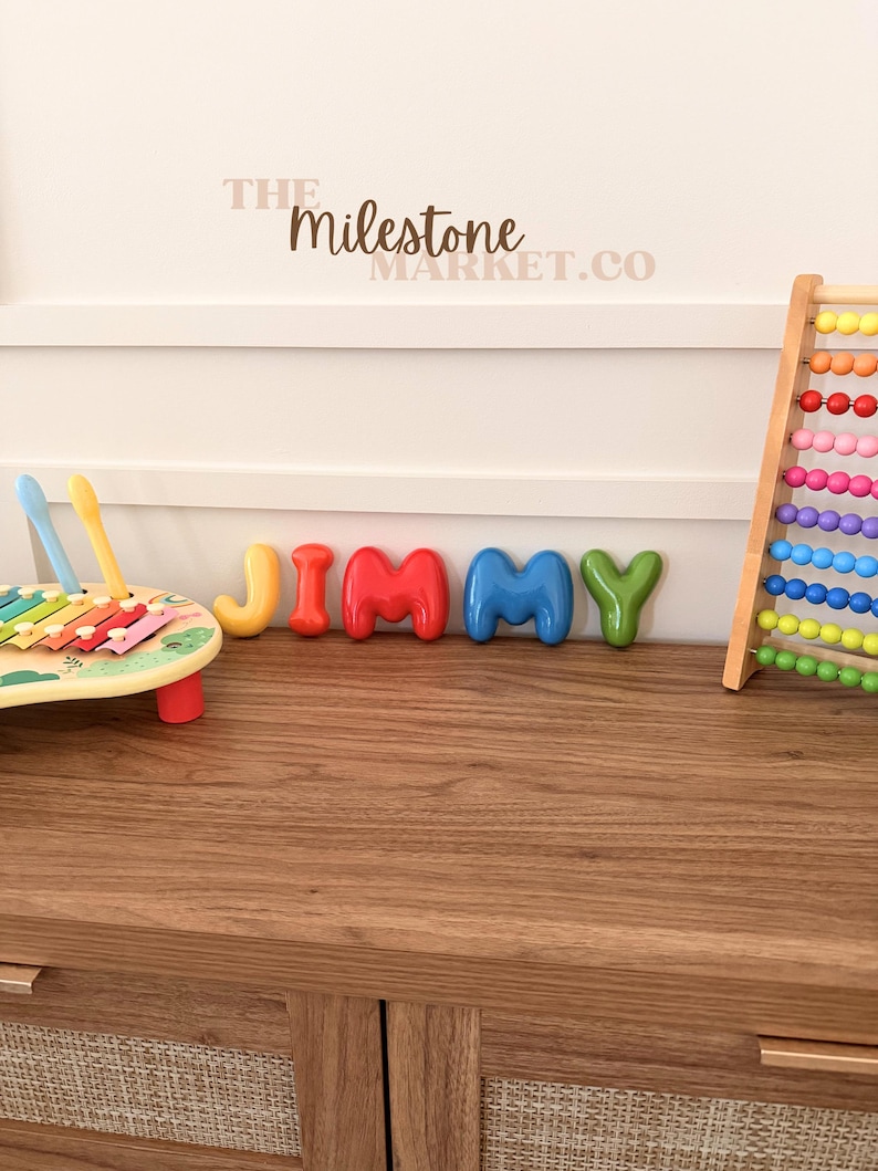 May include: A set of colourful, rounded, three-dimensional letters spelling the name JIMMY, positioned between a wooden xylophone with two mallets and a vertical abacus with rows of multi-coloured beads. The text THE MILESTONE MARKET.CO is visible.