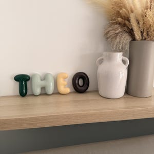 May include: A set of five individual, smooth, rounded, three-dimensional letters spelling the name THEO. The letter T is dark green, H is light sage green, E is pale yellow, and O is dark brown. The letters are arranged in a horizontal row.