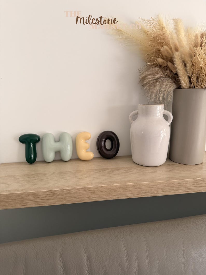 May include: A set of five individual, smooth, rounded, three-dimensional letters spelling the name THEO. The letter T is dark green, H is light sage green, E is pale yellow, and O is dark brown. The letters are arranged in a horizontal row.