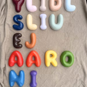 May include: A collection of twenty-one individual, three-dimensional, glossy, balloon-style letters in various colours including red, blue, yellow, green, orange, purple, brown, and pastel shades. The text "THE Milestone MARKET CO" is printed at the bottom.